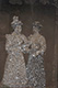 Two ladies with fine dresses and hats, arm in arm. Woman to the right holding a book and parasol in left hand
